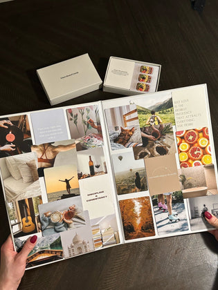  An open vision board book filled with aesthetic collage cards and motivational quotes, alongside a set of vision board prompt cards on a dark wooden table.