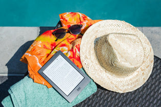  Poolside essentials laid out on a chair: straw hat, sunglasses, orange floral shirt, mint green towel, and a Kobo e-reader displaying text.